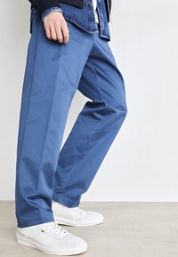 Pantalones azules con una textura suave, corte recto y pliegues delanteros, combinados con zapatillas blancas sobre una superficie neutra.