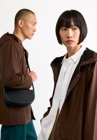 Veste imperméable marron avec une capuche noire, associée à une chemise blanche à boutons. Une personne en arrière-plan porte un sac à bandoulière noir.