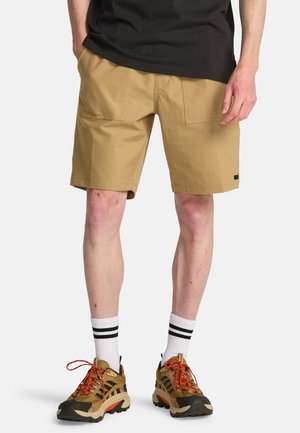 Homme portant un short beige jusqu'aux genoux, des chaussettes blanches à rayures noires et des chaussures de randonnée marron avec des lacets rouges, debout les mains dans les poches.