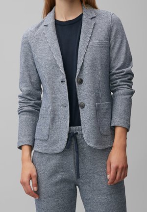 Kvinna som bär en grå och vit rutmönstrad blazer och matchande byxor med dragsko, med en svart tröja under, stående mot en enkel bakgrund.