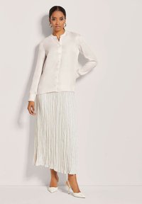 Zijde, lange mouwen shirt in lichtbeige, met een mandelinekraag en voor knopen, gecombineerd met een geplooide, witte maxi-rok en witte hakken.