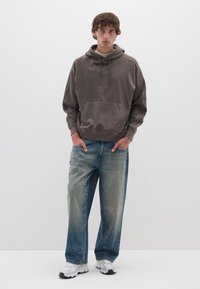 Hoodie marrone oversized con tasca frontale, abbinato a jeans azzurri chiari strappati con gamba larga e un leggero effetto di lavaggio acido.