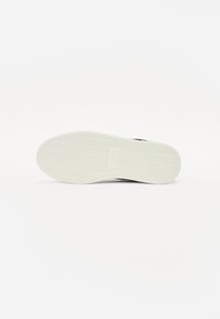 Svart sneaker med en teksturert gummisåle, som har et fiskebensmønster og en lysfarget mellomsåle. Minimalistisk design med subtil merkevare.