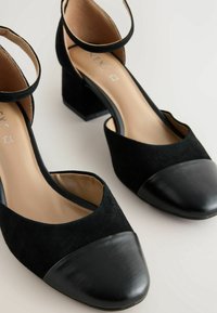 Chaussures à talons carrés en suède noir avec bouts en cuir et brides à la cheville sur fond blanc.
