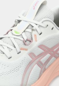 White mesh running shoe with pink and orange accents, textured surface, padded collar, and a prominent brand logo on the tongue.