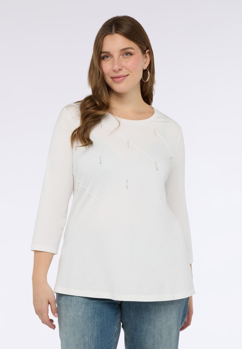 Kobieta ubrana w białą bluzkę z długim rękawem, ozdobioną wzorem w szew diamentowy i małymi wiszącymi koralikami, skomponowaną z niebieskimi dżinsami.