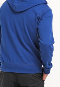 Persona che indossa una felpa con cappuccio blu e pantaloni neri, in piedi con le spalle rivolte verso l'osservatore e una mano in tasca.