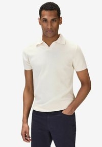 Krämfärgad kortärmad polo med v-ringning, med en texturerad tyg och en åtsittande design, kombinerad med mörkblå byxor.