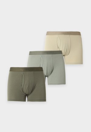 Homme torse nu debout pieds nus, portant un boxer ajusté vert clair, posé avec une main derrière le dos sur un fond uni clair.