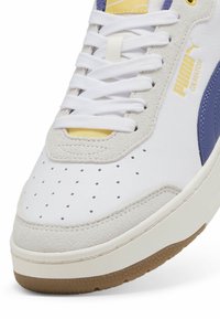 White sneaker with purple and yellow accents, featuring perforated design, smooth leather and suede textures, and rubber sole with grip pattern.