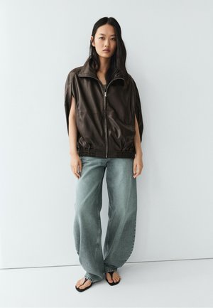 Donna in piedi contro una parete bianca che indossa una giacca marrone scuro larga con zip, jeans larghi scoloriti e sandali neri.