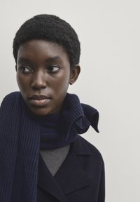 Granatowa, prążkowana szalik otula szyję, w połączeniu z szarym topem i ciemnym płaszczem. Widoczna jest miękka tekstura oraz wyraźny detal związania.