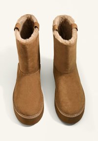 Brown suede ankle boots with a soft, faux fur lining. Features a round toe, flat sole, and two pull tabs at the top for easy wear.