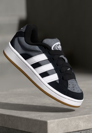 Baskets Adidas noires et grises avec des bandes blanches et une semelle en gomme, présentées en l'air au-dessus d'une surface en béton sous un éclairage doux.