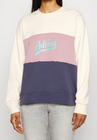 Sudadera de algodón con bloques de color crema, rosa y azul marino, con el logo "Adidas" en turquesa en el pecho. Cuello redondo y puños acanalados.