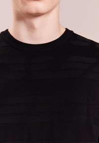 Svart stickad tröja med ett texturerat mönster över tyget, med rund halsringning och en åtsittande design.