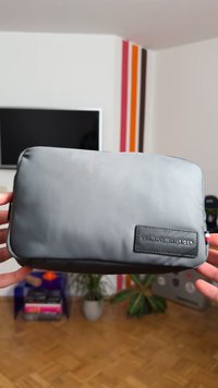 Gray rectangular pouch made of smooth fabric, featuring a dark label with the brand name. Zippered closure, with minimalistic design.