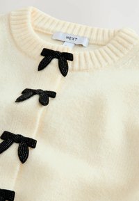 Cream sweater with ribbed neckline, featuring three decorative black bows made of fabric at the front. Soft texture with a subtle pattern.