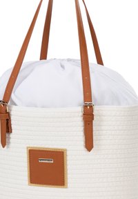 Witte geweven totebag met bruine leren handvatten en gouden hardware. Heeft een trekkoordsluiting onder een witte stoffen bovenkant en een merklabel.