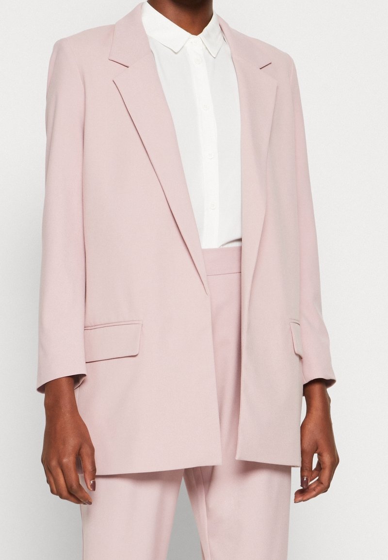 Lichtroze gestructureerd blazer met ingesneden revers, twee voorzakken en lange mouwen, gedragen over een wit overhemd met knoopsluiting.