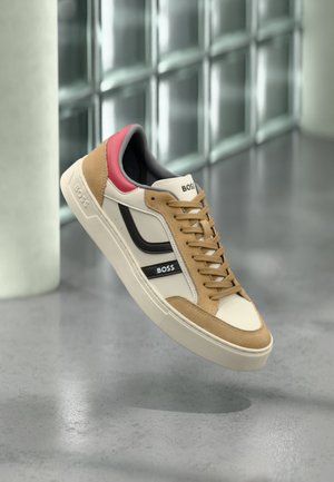 Sneaker alla moda in beige, marrone chiaro, nero e rosso con logo "BOSS", che fluttua sopra un pavimento grigio con pareti di blocchi di vetro sullo sfondo.