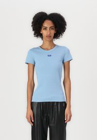 Camiseta de algodón de rayas azul claro con cuello redondo y mangas cortas. Presenta un pequeño logo de la marca en azul en el pecho. Combinada con pantalones negros.