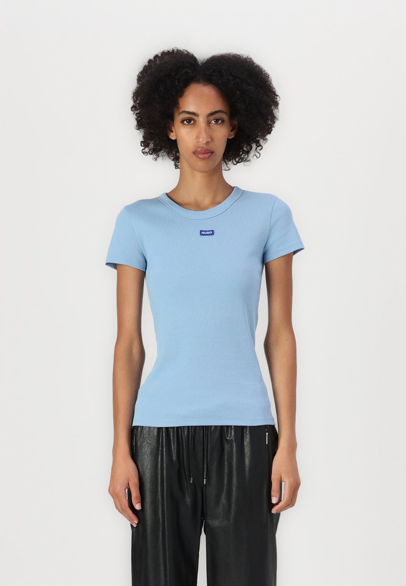 Camiseta de algodón de rayas azul claro con cuello redondo y mangas cortas. Presenta un pequeño logo de la marca en azul en el pecho. Combinada con pantalones negros.