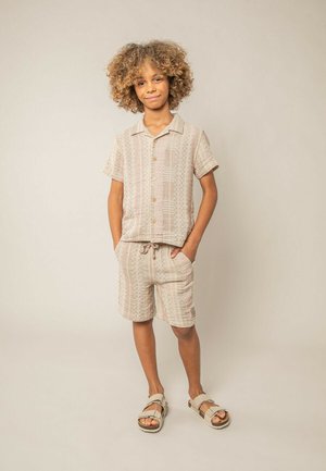 Garçon aux cheveux bouclés portant une chemise à manches courtes beige avec un motif, un short assorti et des sandales beiges, debout les mains dans les poches devant un fond uni.