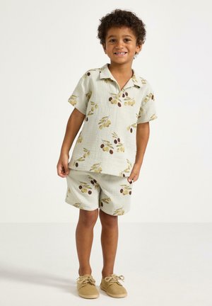 Smilende barn iført cremefarvet kortærmet skjorte og shorts med print af olivengrene samt beige snøresko, stående på ensfarvet baggrund.