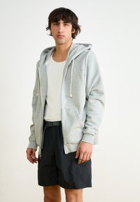 Pelēka zip-up kapuce ar iespējamajiem auklīšu, sānu kabatām un ribotām aprocēm, uzvilkta virs baltā krekla bez piedurknēm un melnām šortiem. Mīksta auduma struktūra.