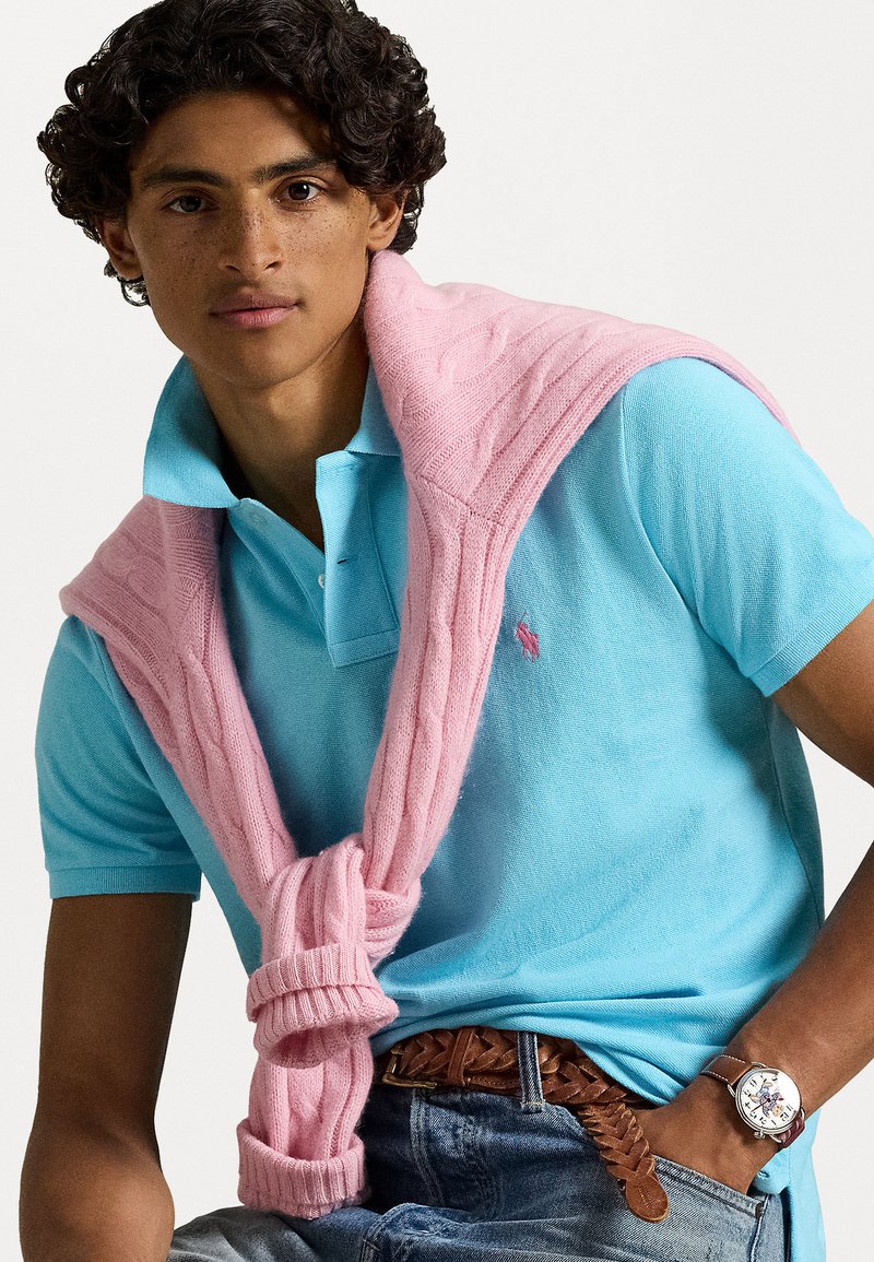 Helles blaues Polohemd mit einem rosa kabelgestrickten Pullover um die Schultern; Jeans; brauner geflochtener Ledergürtel; runde Armbanduhr.