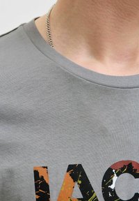 Harmaa puuvillapaita, jossa on pyöreä kaula-aukko. Edessä värikäs roiskeprintti oranssilla, mustalla ja vihreällä. Kaulaketju näkyvissä kaula-aukossa.