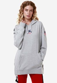 Felpa grigia con tasca davanti, presenta un logo "US" con stelle e una patch della bandiera americana su una manica. Tessuto morbido, vestibilità rilassata.