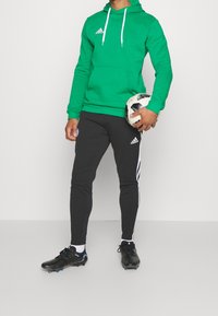 Sudadera verde con cordones blancos, pantalones negros ajustados con franjas laterales blancas y botas de fútbol negras, sosteniendo un balón de fútbol en blanco y negro.