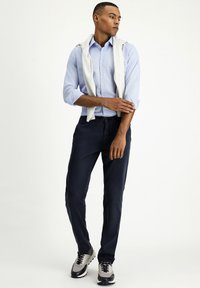 Lichtblauw gestreept overhemd met opgerolde mouwen, marineblauwe broek en grijze sneakers. Trui nonchalant over de schouders gedrapeerd.