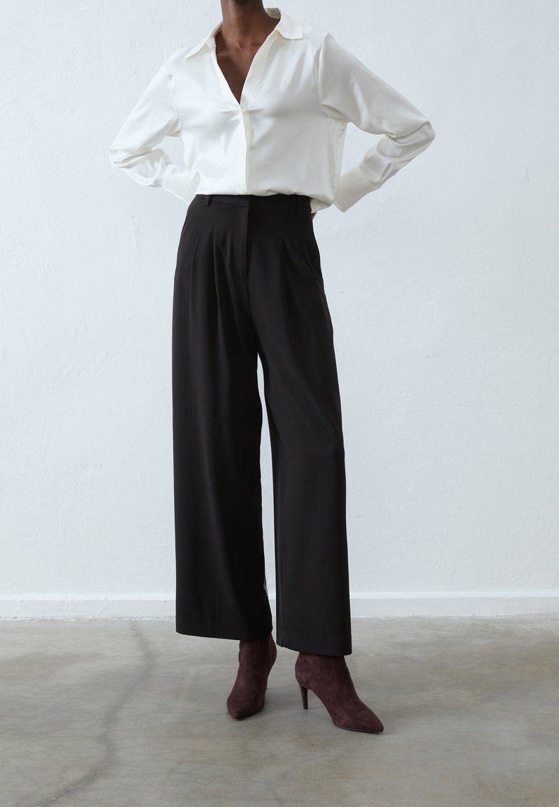 Personne portant une chemise blanche boutonnée, un pantalon noir à jambes larges et des bottines à talons hauts pointues de couleur sombre, debout sur un sol en béton.