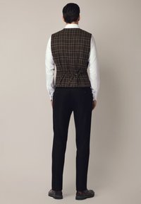 Brązowa kamizelka w kratę o dopasowanym kroju, z wiązaniem w talii i białą koszulą z długim rękawem, w połączeniu z czarnymi spodniami i ciemnymi butami.