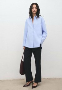 Camicia azzurro chiaro con taschino sul petto, abbinata a pantaloni neri a gamba larga e tacchi a punta bordeaux, completata da una borsa coordinata.