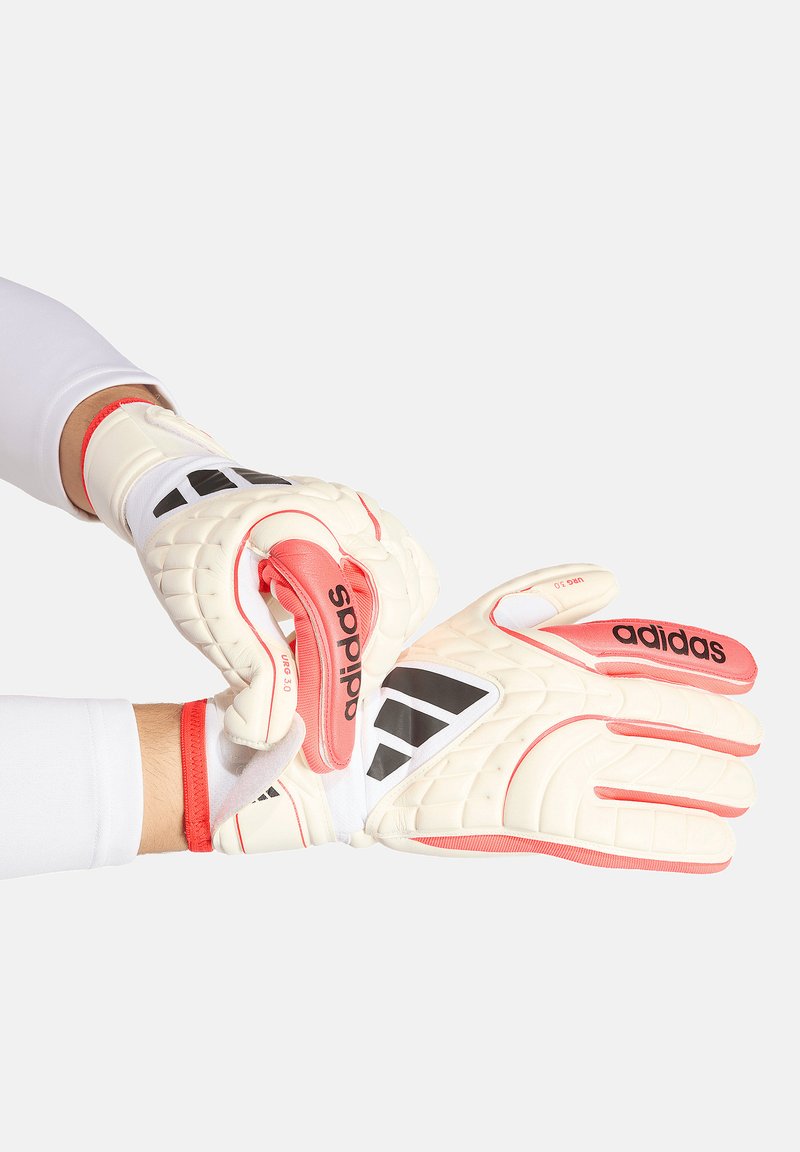 Mains portant des gants de gardien Adidas blancs et corail, fixant la sangle du poignet sur une manche blanche.
