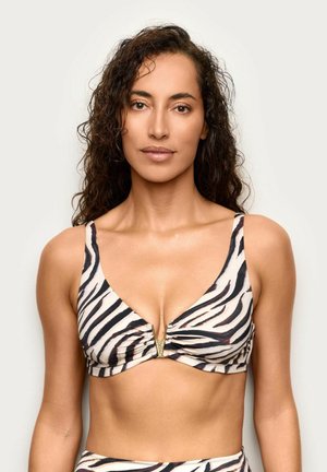 Donna con capelli ricci che indossa un top bikini a fantasia zebra con dettaglio dorato a forma di V, in piedi davanti a uno sfondo bianco uniforme.