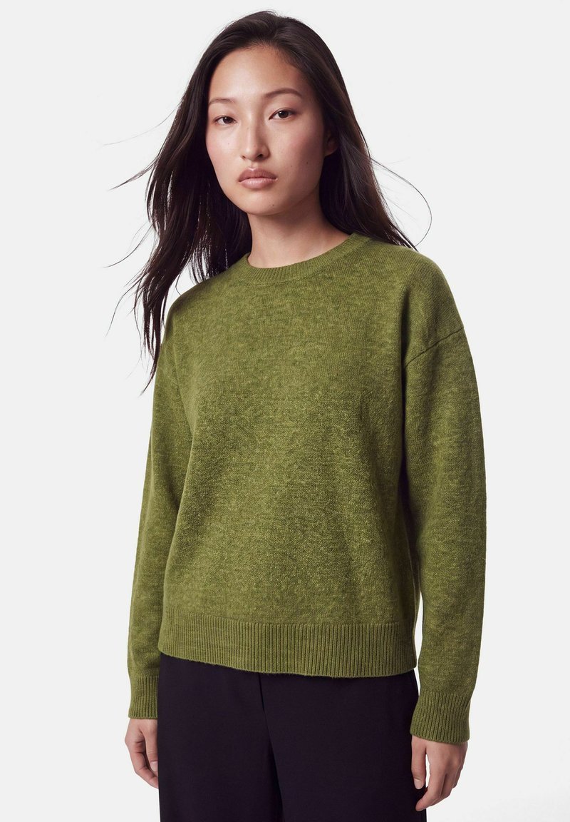 Jeune femme aux longs cheveux foncés, portant un pull en tricot vert et un pantalon noir, debout devant un fond clair uni.