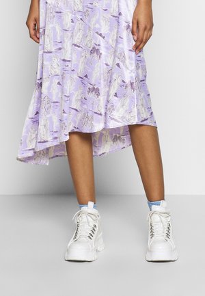 Nogi w liliowej spódnicy z białymi nadrukami klasycznych rzeźb, zestawione z białymi masywnymi butami na platformie i jasnoniebieskimi skarpetkami.