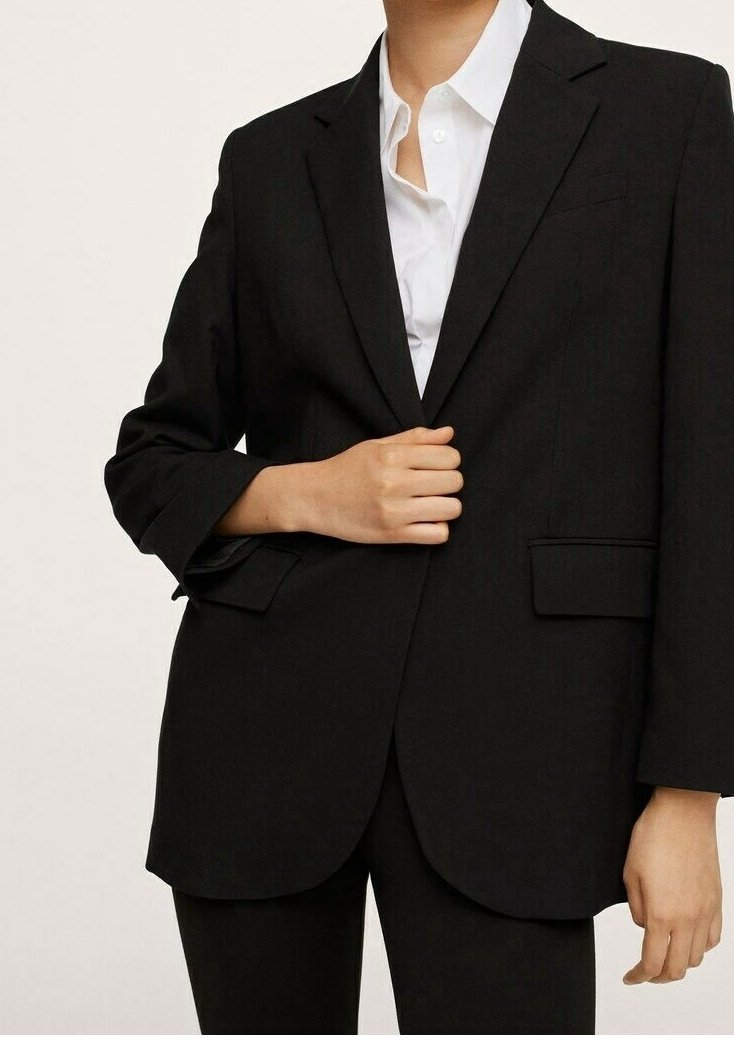 Personne portant un blazer noir sur une chemise blanche boutonnée, une main ajustant le devant du blazer, sur un fond uni.