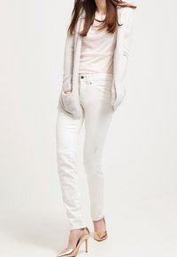 Jeans slim fit bianchi con leggeri strappi, abbinati a una maglietta rosa chiaro e un cardigan grigio strutturato. Accessorio: tacchi alti dorati lucidi.
