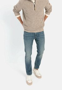 Beige Strickpullover mit Reißverschlusskragen, kombiniert mit hellblauen Slim-Fit-Jeans und weißen Sneakers mit beigen Akzenten.
