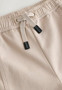 Shorts beiges avec taille élastique, cordon de serrage ajustable et petites étiquettes de marque noires. Le matériau présente une finition lisse et texturée.