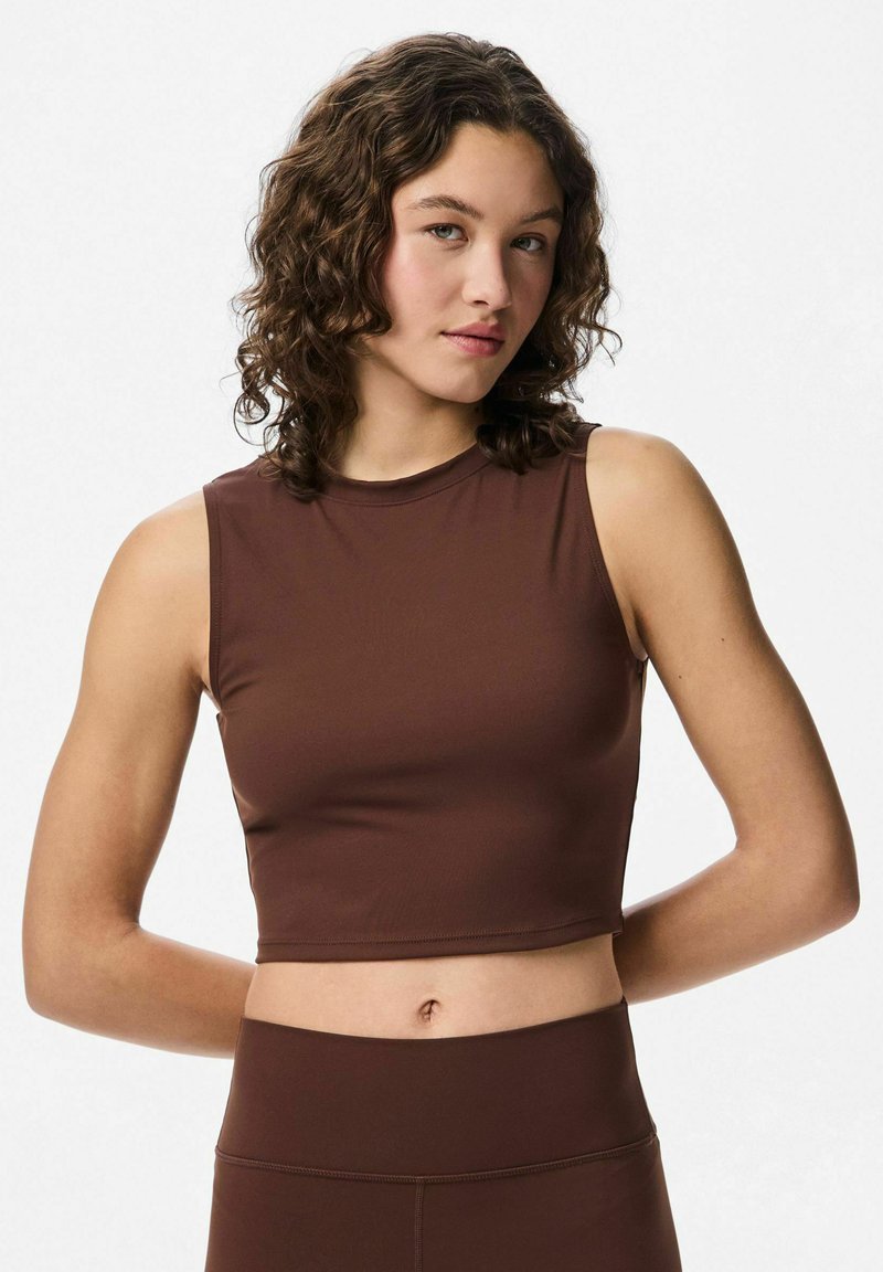 Jeune femme aux cheveux bouclés portant un crop top sans manches de couleur marron et un legging taille haute assorti, se tenant les bras derrière le dos.