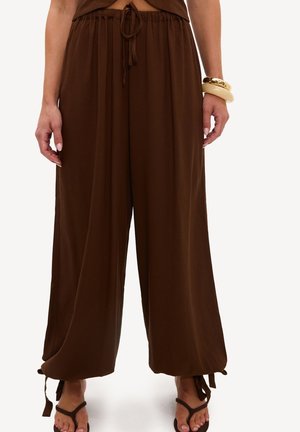 Personne portant un pantalon ample marron foncé avec des cordons aux chevilles et des sandales marron foncé, également des bracelets au poignet droit.
