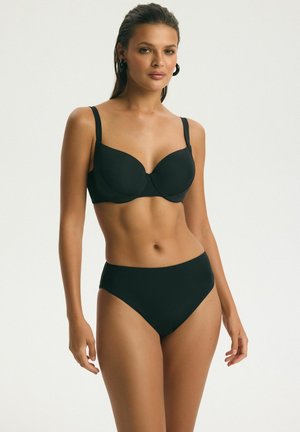 Kobieta stojąca, ubrana w czarny biustonosz z fiszbinami i dopasowane majtki z wysokim stanem, o neutralnym wyrazie twarzy i długich ciemnych włosach zaczesanych do tyłu.