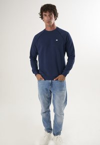 Granatowa bluza crewneck z raglanowymi rękawami, prążkowanymi mankietami i małym białym logo. Zestawiona z jasnoniebieskimi prostymi dżinsami i białymi trampkami.